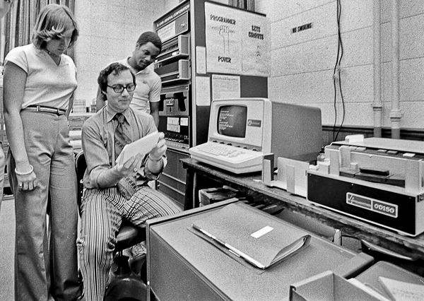 Black and white photo of a computer lab with three individuals at work.