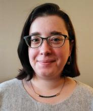 Sari, a white person with shoulder-length, side-parted brown hair and glasses, smiles slightly and looks at the camera. The images is a portrait and they are wearing a beige sweater and a necklace