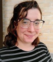 Sari, a white person with wavy-curly brown hair and blue rimmed glasses wearing a black and white striped top, sits in front of a yellow brick wall and smiles, closed mouth, at the camera.